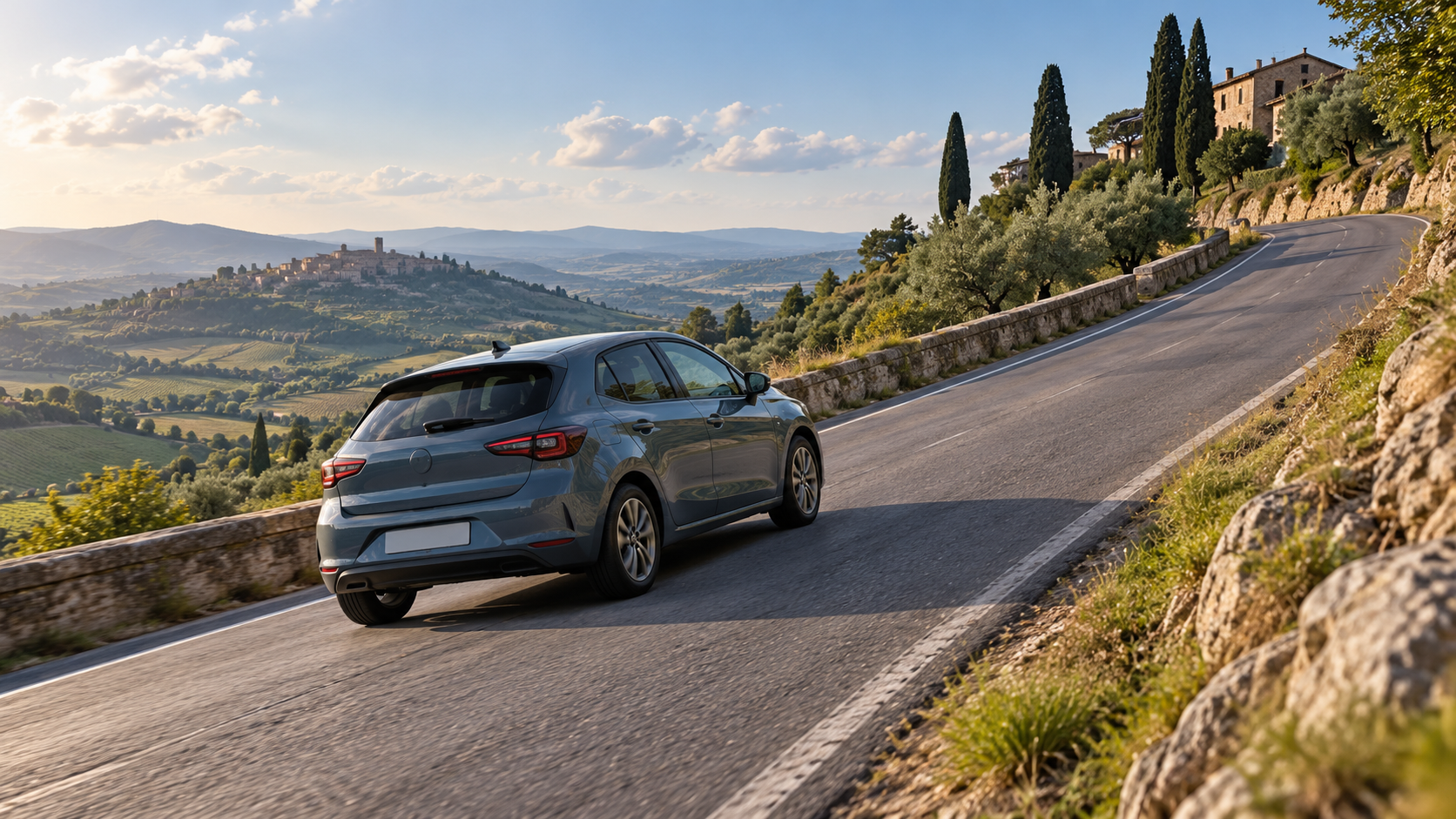 Auto grigio blu su una strada in salita tra le colline, con un borgo sullo sfondo